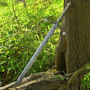 Op de afbeelding: Een lang, zilveren zwaard met ingewikkelde geëtste patronen op het lemmet en de handgreep. Het zwaard heeft een pareerstang met decoratieve elementen en leunt tegen een boomstam in een natuurlijke omgeving. Het ontwerp suggereert een historisch of fantasiethema.