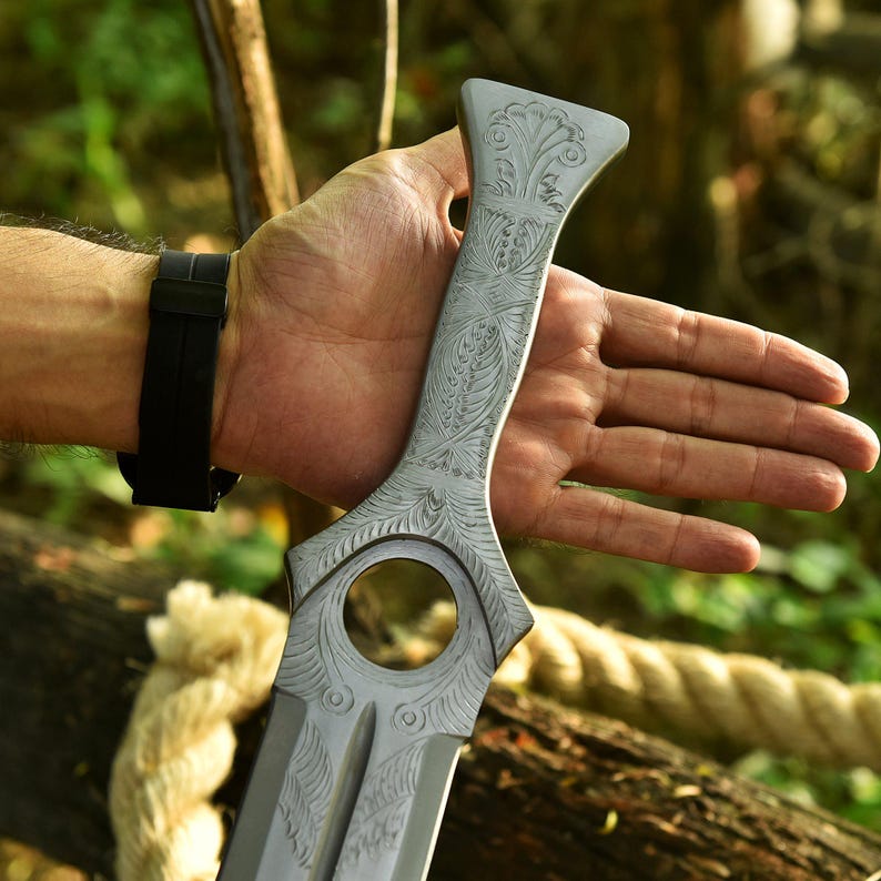 May include: A silver-colored sword with intricate floral engravings. The blade has a unique shape with a circular cutout near the handle. The handle also features detailed carvings. The sword is held against a natural, blurred background.