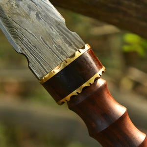 May include: Close-up of a Damascus steel knife blade with a dark wood handle. The blade has a wavy pattern, and the handle features a brass accent. The knife is designed for cutting.