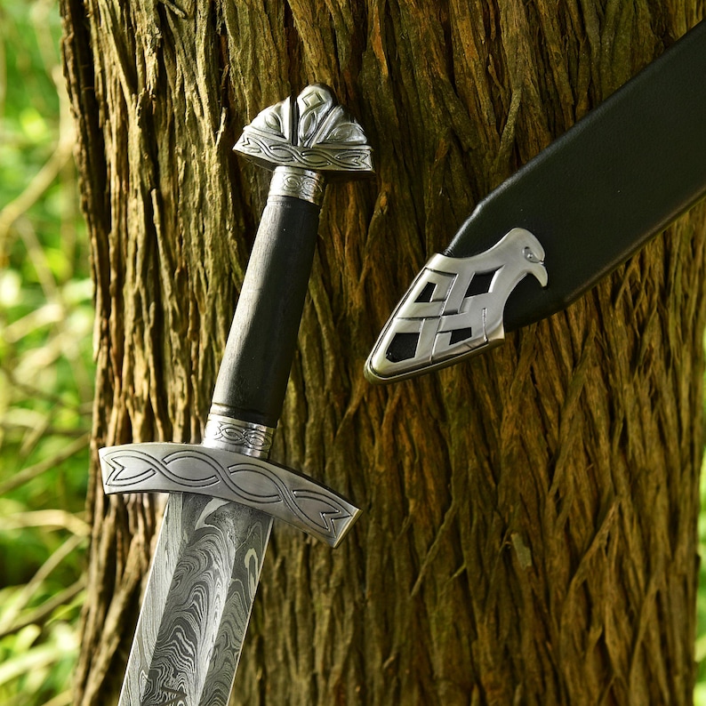 May include: A detailed image of a sword with a patterned blade and ornate silver hilt. The handle is black, and the scabbard is visible. The sword is leaning against a tree trunk, showcasing the craftsmanship.