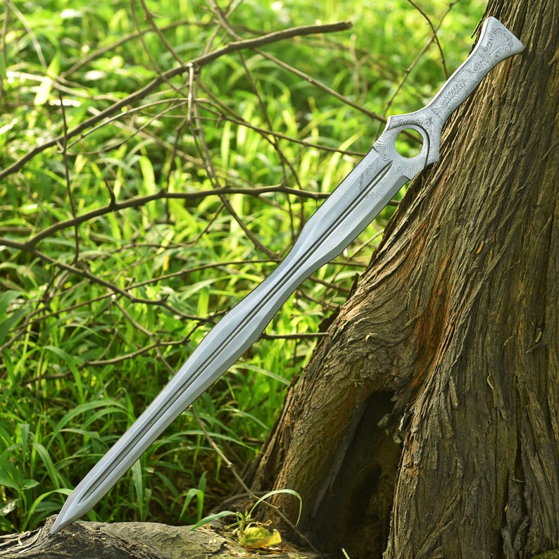 May include: A silver-colored sword with an ornate handle and a long, pointed blade. The sword is leaning against a tree trunk, with green foliage in the background. The handle features a circular design and engraved patterns.