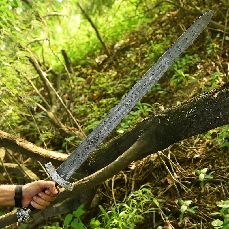 May include: A long, ornate sword with a patterned blade and a decorative handle. The sword is held against a backdrop of green foliage and a fallen tree branch. The blade has text etched into it.