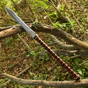 May include: A long, ornate spear with a patterned, silver-colored blade. The handle is a dark brown color with a ridged design, and the base is accented with gold-colored metal. The spear is displayed against a backdrop of natural wood and foliage.