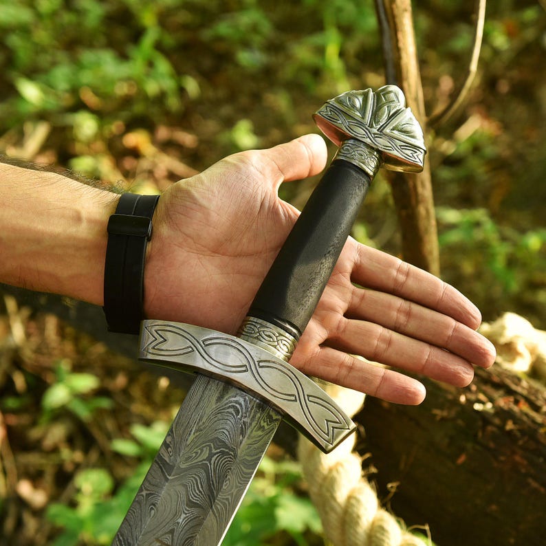 May include: A detailed image of a sword with a patterned blade and ornate silver crossguard and pommel. The handle is black, and the blade has a unique, swirling pattern. The sword is held in a hand, with a black watch band visible on the wrist.