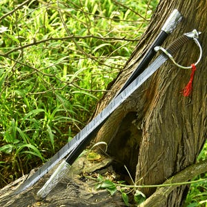 May include: A decorative sword with a patterned blade and a silver hilt rests against a tree trunk. The sword has a brown handle and a red tassel. The scabbard is black with silver accents.