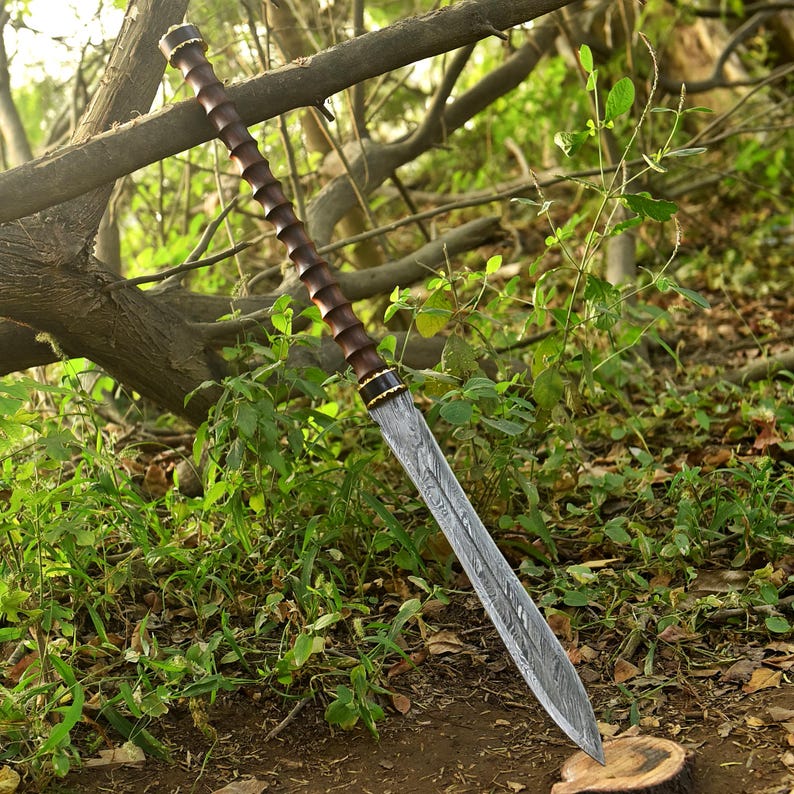 May include: A long, ornate sword with a patterned blade and a dark wooden handle. The handle has a spiral design and is accented with gold-colored bands. The sword is resting against a tree branch in a natural outdoor setting.