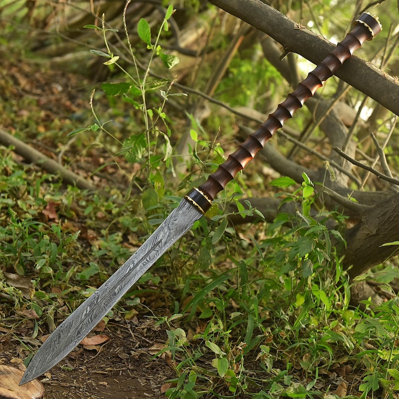 May include: A long, ornate sword with a patterned blade and a dark wooden handle. The handle has a ridged design with gold accents. The sword is set against a backdrop of green foliage and tree branches.