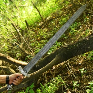 May include: A long, ornate sword with a patterned blade and a decorative handle. The sword is held against a backdrop of green foliage and a fallen tree branch. The blade has text etched into it.