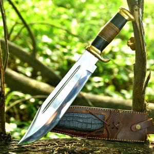 May include: A large, polished steel hunting knife with a brown leather and black handle, accented with gold-colored metal. The knife rests on a log with a brown leather sheath with a crocodile skin pattern.