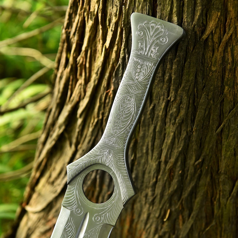 May include: Close-up of a silver-colored, ornate sword blade with intricate engraved floral and geometric patterns. The blade features a circular opening near the center and rests against a textured tree trunk, suggesting a fantasy or historical theme.