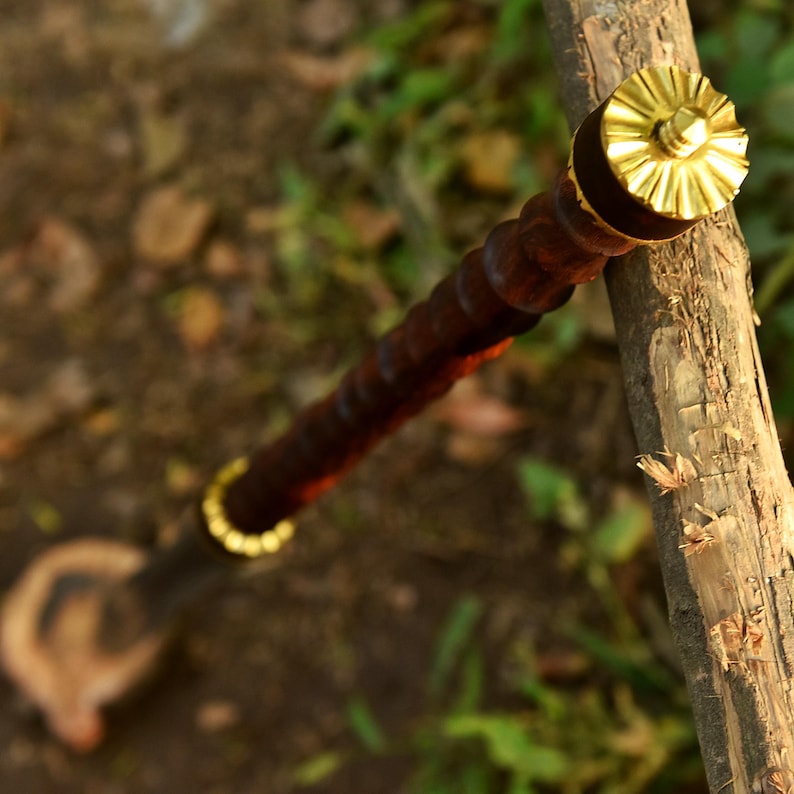 May include: A dark brown wooden walking stick with a gold-colored metal top and bottom ring. The top has a decorative, sunburst-like design. The stick rests on a piece of wood, with a blurred green and brown background.