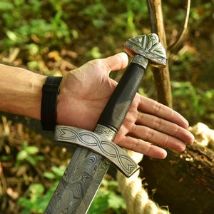 May include: A detailed image of a sword with a patterned blade and ornate silver crossguard and pommel. The handle is black, and the sword is held in a hand with a black watch.