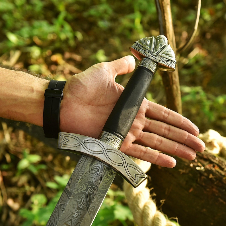 May include: A detailed image of a sword with a patterned blade and ornate silver crossguard and pommel. The handle is black, and the sword is held in a hand with a black watch.