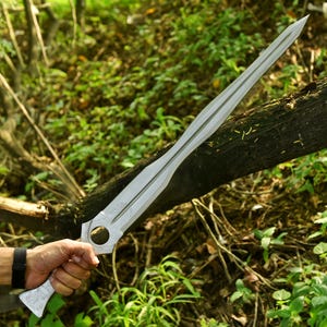 May include: A silver-colored sword with a long, double-edged blade. The hilt features a circular opening and decorative engravings. The sword is held in a hand with a black watch. The background is a natural outdoor setting with green foliage.