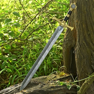 Op de afbeelding: Een lang, sierlijk zwaard met een patroon op het lemmet en een goudkleurige pareerstang. De handgreep is zwart met gouden details. Het zwaard leunt tegen een boomstam, met groene bladeren op de achtergrond.