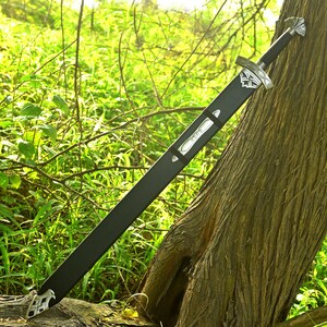 May include: A long, black sword with a silver hilt and accents, resting against a tree. The sword is in a black sheath with silver details. The background features green foliage and sunlight.