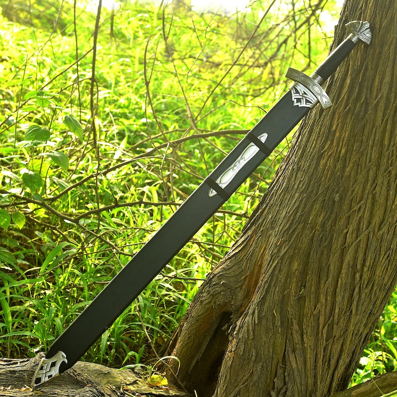 May include: A long, black sword with a silver hilt and accents, resting against a tree. The sword is in a black sheath with silver details. The background features green foliage and sunlight.