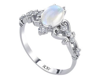 May include: A silver ring with an oval, iridescent gemstone at the center, surrounded by smaller, clear gemstones and decorative filigree. The band is thin and polished, with the word "ROSY" engraved on the inside.