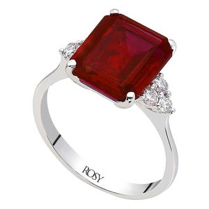 Può includere: Un anello d'argento con una grande gemma rettangolare rosso scuro come elemento centrale. Piccole pietre chiare affiancano la pietra principale. La fascia dell'anello è liscia e lucida, con la scritta "ROSY" incisa all'interno.