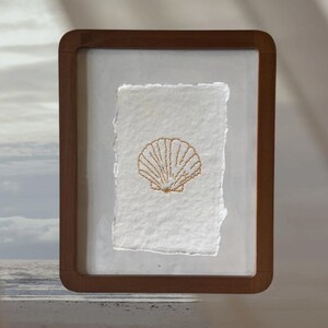 Op de afbeelding: Een ingelijst kunstwerk met een gouden schelpmotief op getextureerd wit papier. De lijst is van donkerbruin hout. De achtergrond toont een strandscène. Afmetingen: ongeveer 20 cm x 30 cm.