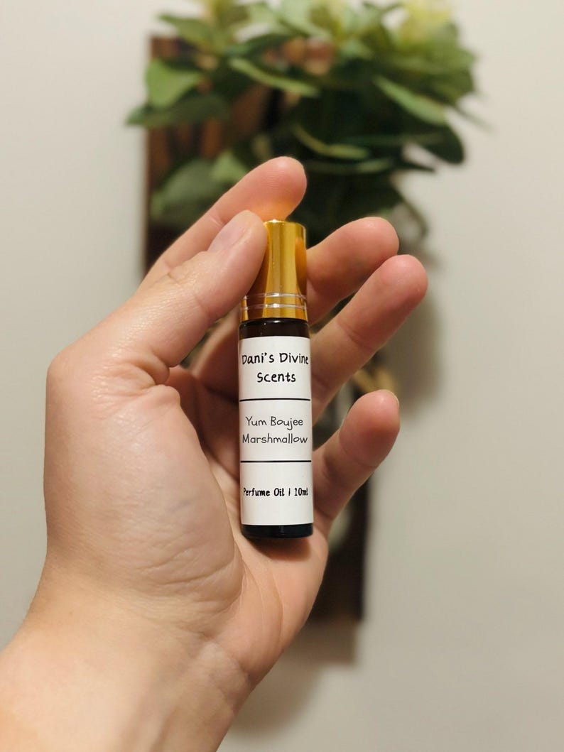 Op de afbeelding: Een kleine parfumolieflacon met een gouden dop en een zwarte basis wordt in een hand gehouden. Het etiket luidt "Dani's Divine Scents" en "Yum Boujee Marshmallow". De fles bevat 10 ml parfumolie.