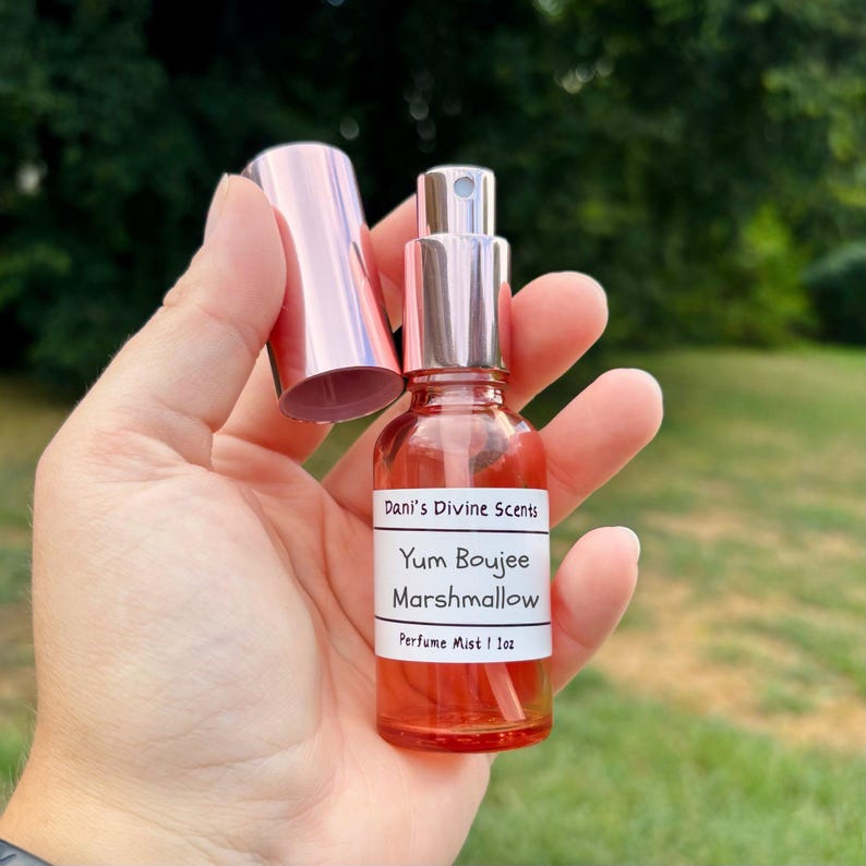 Op de afbeelding: Een kleine, heldere glazen parfumfles met een roze metalen dop en verstuiver. Het etiket luidt "Dani's Divine Scents Yum Boujee Marshmallow Perfume Mist 1 oz.". De fles bevat een rood-oranje vloeistof.