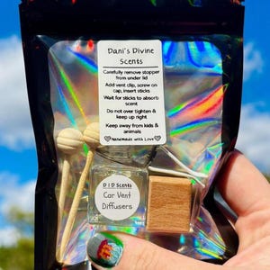 May include: A clear glass car vent diffuser with wooden sticks and a wooden block, packaged in a black holographic bag. The label reads "Dani's Divine Scents" and "Car Vent Diffusers."