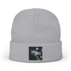 May include: A light gray beanie with a square embroidered patch. The patch features a stylized airplane design in shades of green, purple, and white against a black background. The beanie is a classic, cuffed style.