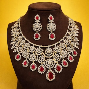 Puede incluir: Un conjunto de collar y pendientes dorados con piedras preciosas claras y rojas. El collar presenta un diseño floral con piedras rojas en forma de lágrima colgantes. Los pendientes a juego tienen un diseño similar. Las joyas se exhiben en un maniquí marrón.