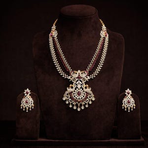 Puede incluir: Un conjunto de collar y pendientes dorados, exhibido sobre un busto marrón. El collar presenta múltiples hilos con detalles blancos y burdeos, y un colgante central con diseño de pavo real. Los pendientes reflejan el diseño del colgante.