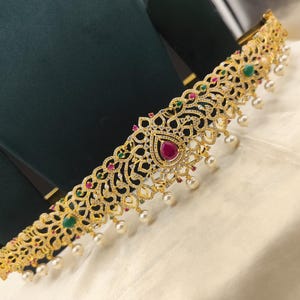 Könnte beinhalten: Goldfarbener Taillengürtel mit aufwendigem Filigranmuster. Der Gürtel hat einen tropfenförmigen Rubin in der Mitte, umgeben von kleineren grünen und roten Edelsteinen. Weiße Perltropfen hängen am unteren Rand und erzeugen einen eleganten, verzierten Look.
