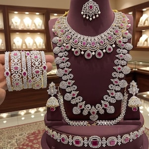 May include: A complete jewelry set featuring a necklace, earrings, bracelet, and headpiece. The set is displayed on a purple mannequin. The jewelry is silver-toned with clear and red gemstones. The background shows jewelry displays.