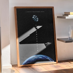 Può includere: Un poster incorniciato con sfondo scuro e la parola "Interstellar" in bianco. Il poster presenta un pianeta blu, un ammasso di stelle e un fascio di luce. La cornice è in legno.