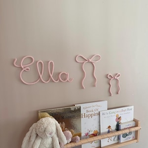 May include: A pink rope name spelling "Ella" and two bow decorations are mounted on a pale pink wall. A wooden bookshelf below displays children's books and soft toys, suggesting a nursery or child's bedroom.