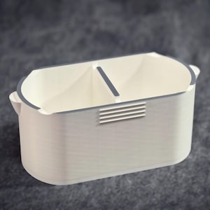 Puede incluir: Un organizador rectangular blanco con un interior dividido. El organizador tiene un borde gris y un pequeño detalle de ventilación rectangular en la parte delantera. El diseño es minimalista y moderno, adecuado para la organización de escritorio.