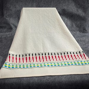 Op de afbeelding: Crèmekleurig textiel met een decoratieve rand. De rand heeft een herhalend patroon van rode en zwarte verticale lijnen, met blauwe en gele accenten. Het textiel is gevouwen, waardoor een driehoekige vorm ontstaat.
