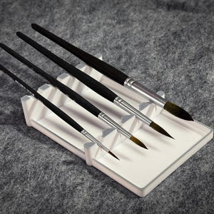 Könnte beinhalten: Ein Set aus fünf Pinseln mit schwarzen Griffen und silbernen Zwingen, die in einem weißen, rechteckigen Pinselhalter liegen. Die Borsten variieren in Form und Größe. Die Pinsel liegen auf einer grauen Oberfläche.