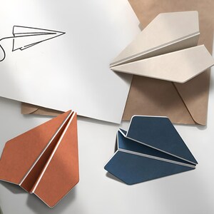 May include: An overhead shot features three paper airplanes in shades of beige, orange, and navy blue, alongside a line drawing of a paper airplane. A brown envelope and a white card are also present, all arranged on a white surface.