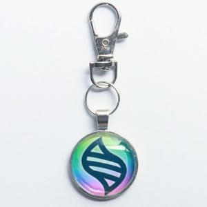 May include: A silver-toned keychain with a round pendant featuring a colorful, iridescent design. The pendant has a blue and white graphic. The keychain includes a clasp and a split ring for attaching keys or accessories.