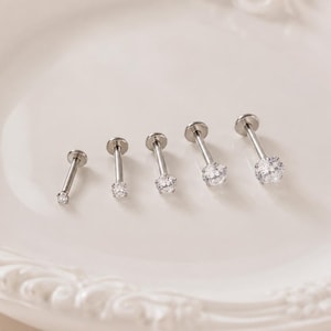 Op de afbeelding: Vijf zilverkleurige labret-piercings op een rij, elk voorzien van een heldere, rondgeslepen edelsteen in een zetting met vier pootjes. De metalen staafjes zijn glad en recht en eindigen in een platte, ronde basis.