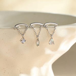 May include: Three silver earrings with delicate chain details and gemstone pendants. Each earring features a different gemstone shape: a square, an oval, and a teardrop. The earrings are displayed in a white ceramic bowl.