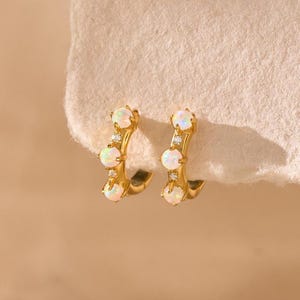May include: A pair of gold-coloured hoop earrings, each featuring three opal-like stones and small clear crystals. The earrings are presented on a textured, off-white surface against a soft, neutral background.
