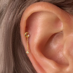 May include: Gold earrings with a crescent moon and star design. The earrings hang from a small stud, with a fine chain connecting the moon and star. The earrings are worn on an ear.