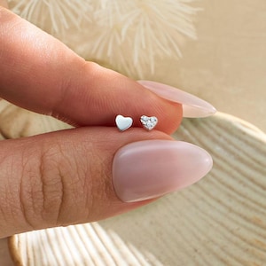May include: Two small silver-toned heart-shaped stud earrings. One earring features a smooth, polished metallic surface, while the other earring is encrusted with tiny, clear, sparkling stones arranged in a heart shape.