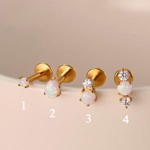 May include: Four gold-coloured stud earrings with white opal and clear crystal accents. The earrings are numbered 1 to 4. The earrings are displayed on a light pink surface.