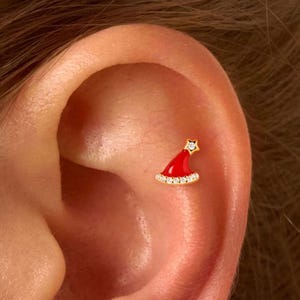May include: Two small, red Santa hat earrings with gold trim and a small star on top. The earrings are set with small, clear stones. The earrings are shown on an ear.