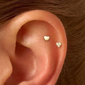 May include: Four small gold heart-shaped stud earrings are worn in the ear. Two earrings are placed in the earlobe, and two are positioned in the upper cartilage. Each earring features a polished gold finish, with some set with tiny clear stones.