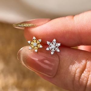 May include: Two small snowflake-shaped stud earrings, one in a gold tone and one in a silver tone, each featuring a central clear stone and small clear stones along the six points of the snowflake design, held between two fingers.