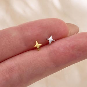 May include: Two small stud earrings shaped like four-pointed stars. One earring is gold-toned and the other is silver-toned. The text Gleamy Day is printed at the bottom of the image. The earrings are positioned on a finger for scale.