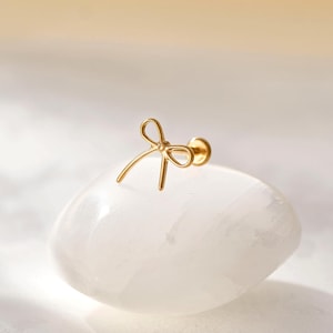 May include: A gold-colored bow-shaped earring stud is displayed on a white, oval-shaped stone. The earring has a delicate, minimalist design. The background is a soft, blurred gradient of beige and white.